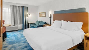 A hotel room with a large bed, a desk, a chair, a sofa, a nightstand, and a wall-mounted lamp.