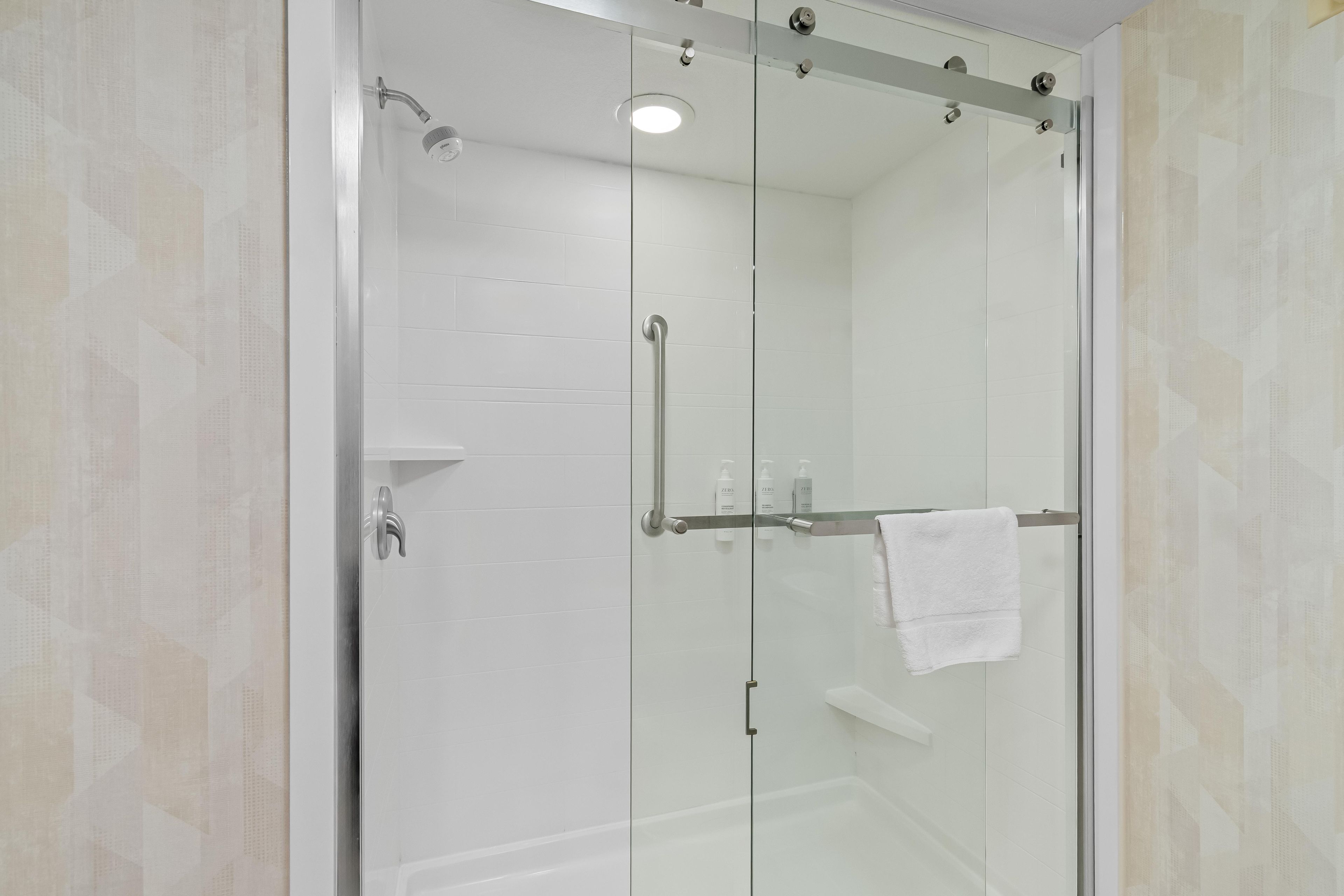 Un box de ducha con paredes de vidrio, una toalla blanca colgada en un perchero, un cabezal de ducha de mano y una puerta de cristal.