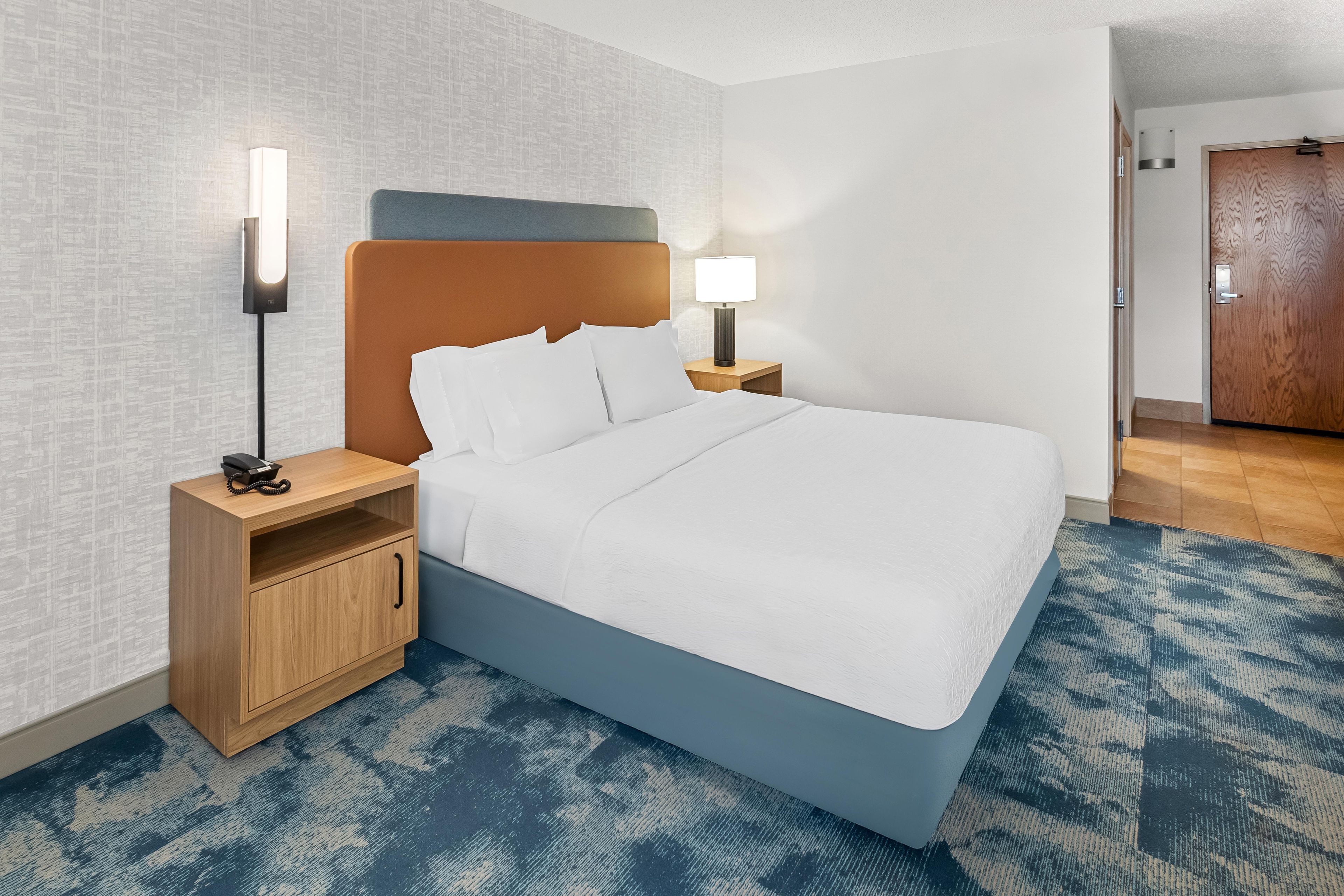 Quarto de hotel com uma cama, mesas de cabeceira, uma luminária, um armário de madeira e uma porta.