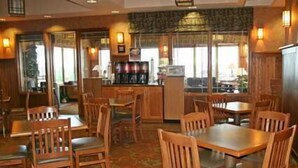 Un restaurante con mesas y sillas de madera, una zona de bar y amplios ventanales que dejan entrar luz natural.
