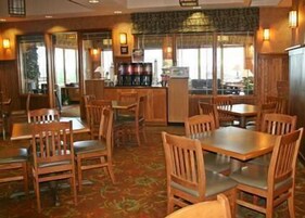 Un restaurante con mesas y sillas de madera, una zona de bar y amplios ventanales que dejan entrar luz natural.