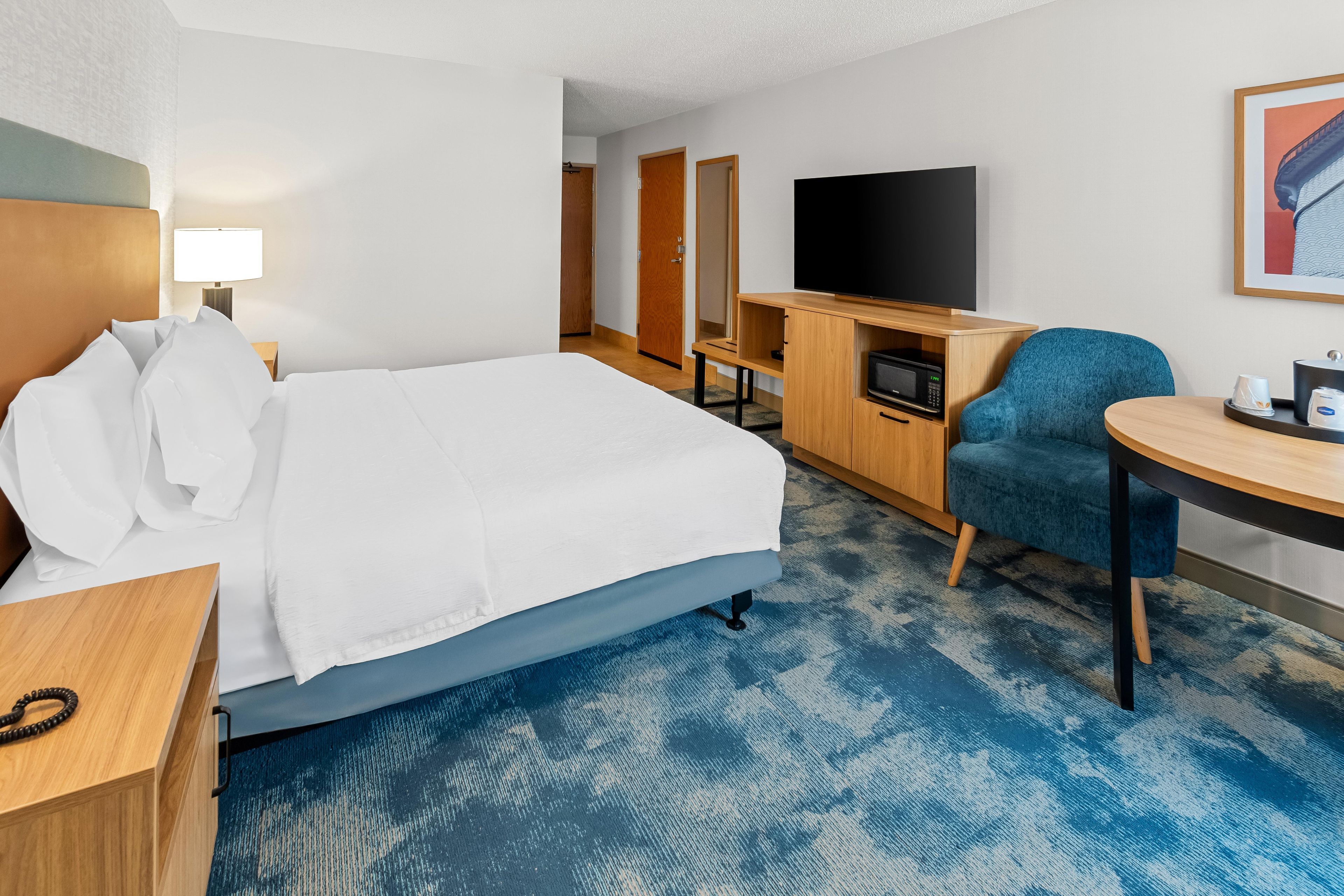 Quarto de hotel com uma cama, escrivaninha, televisão, cadeira e mesa pequena.