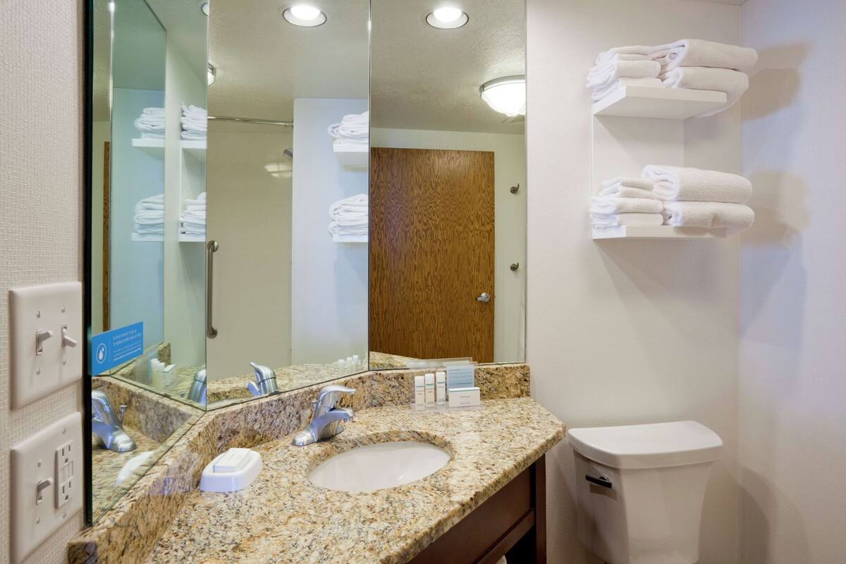 Un baño con lavamanos de granito, espejo, inodoro y toallero con toallas dobladas.