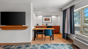 Ett modernt hotellrum med en platt-TV, en matplats med ett träbord och stolar, ett kök med träskåp och ett stort fönster med gardiner.