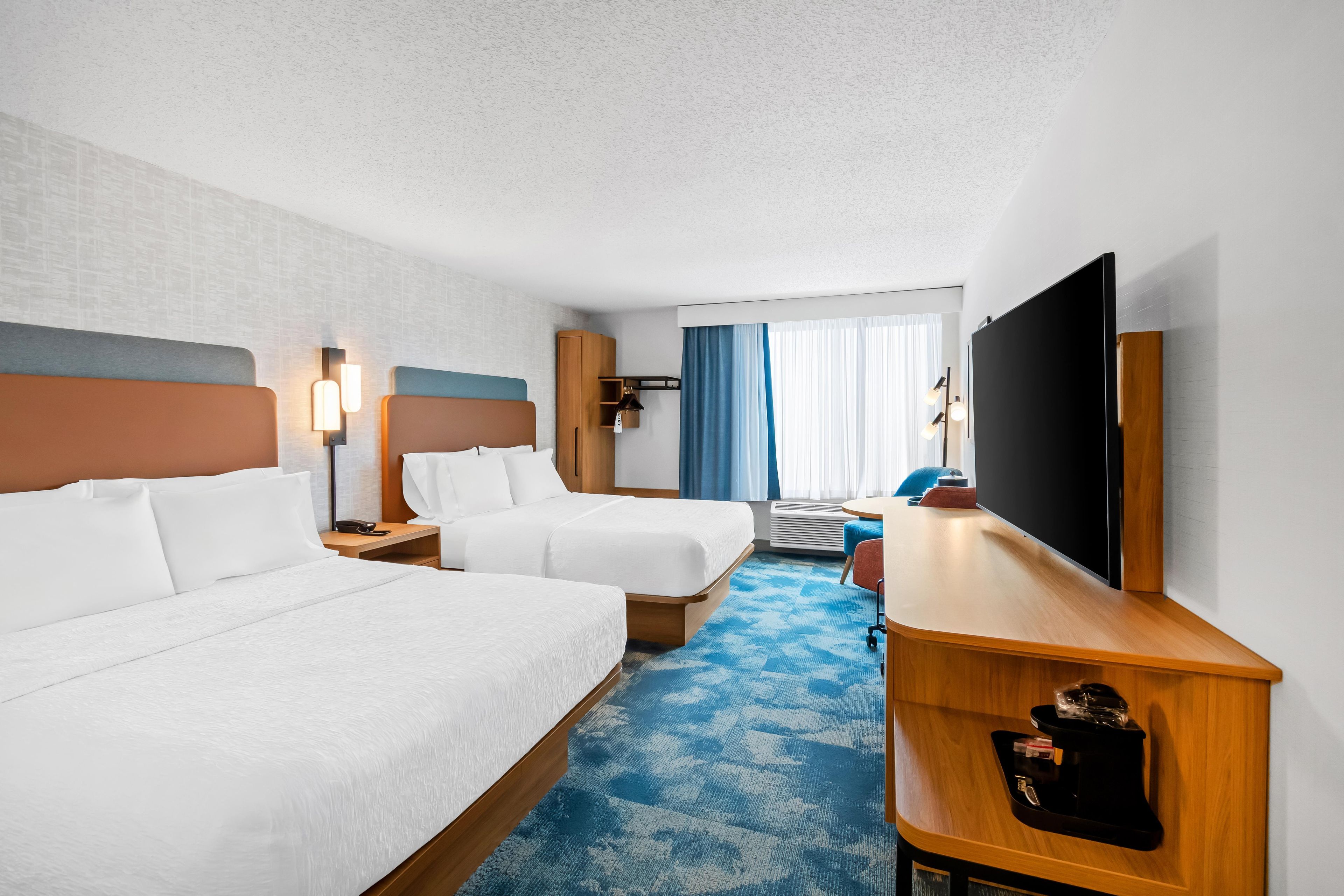 A hotel room with two beds, a desk, a flat-screen TV, a desk lamp, and a window with curtains.