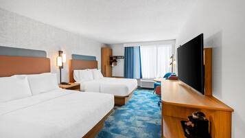 A hotel room with two beds, a desk, a flat-screen TV, a desk lamp, and a window with curtains.