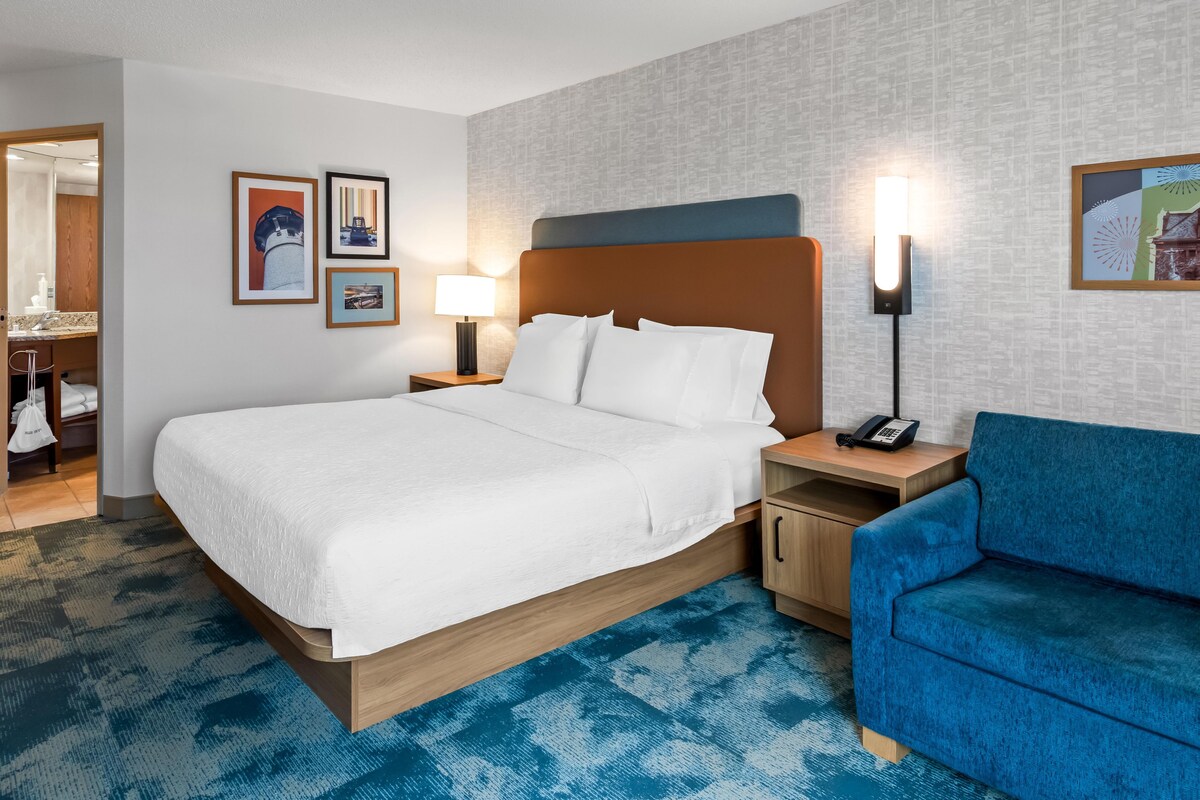 A hotel room with a large bed, a blue sofa, a bedside table with a lamp, and a wall-mounted phone.