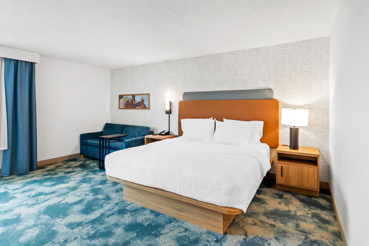 A hotel room with a bed, a desk, a chair, a nightstand, a lamp, and a painting on the wall.