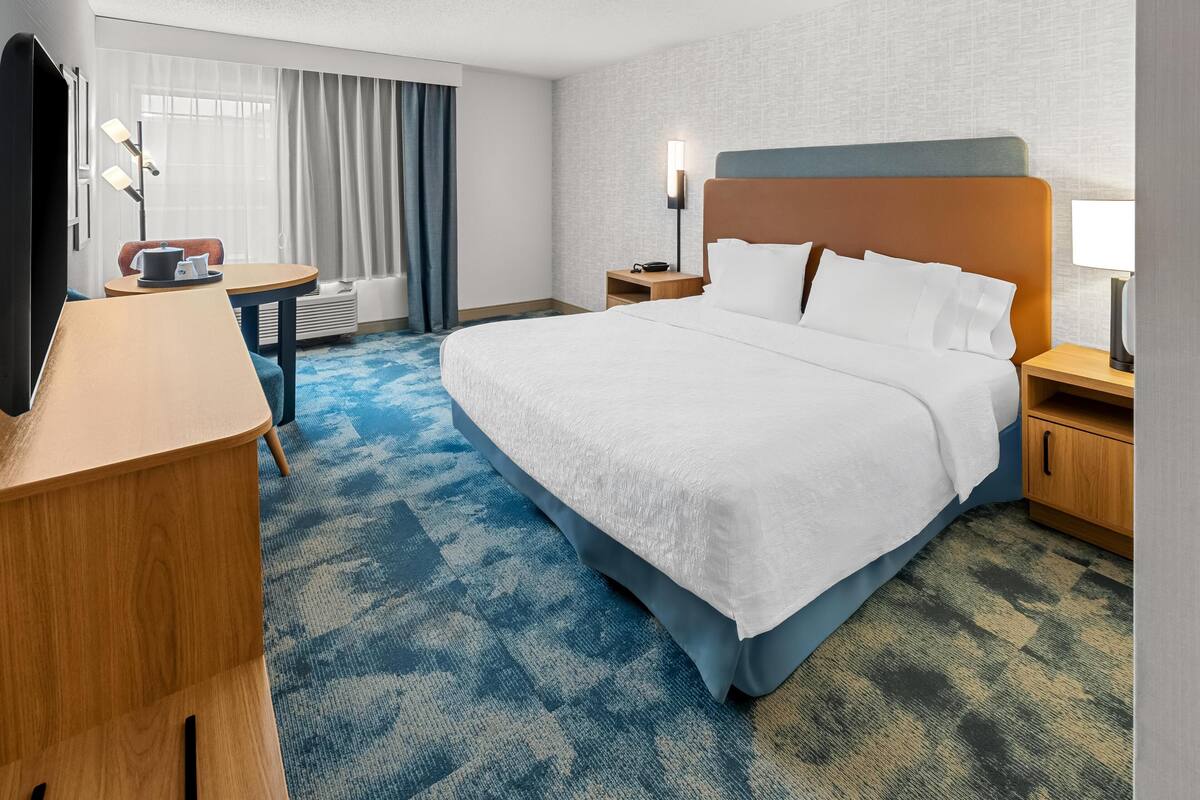 A hotel room with a large bed, a desk, a chair, a small table, and a window with curtains.