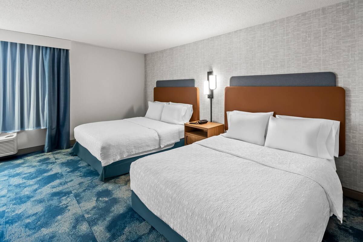 A hotel room with two beds, a blue carpet, a window with curtains, and a wall-mounted lamp.