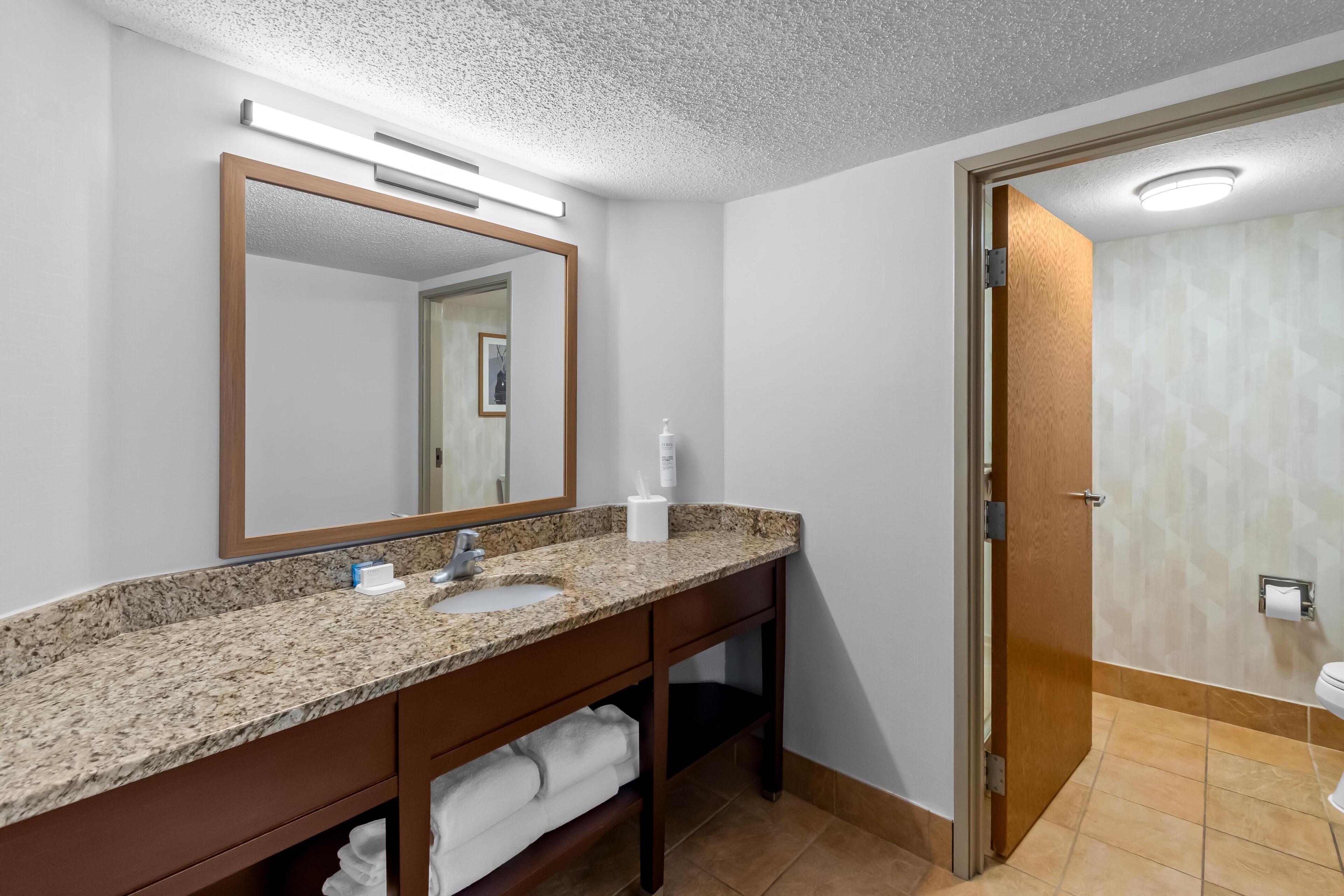 Un baño con encimera de granito, lavamanos, espejo y zona de ducha.