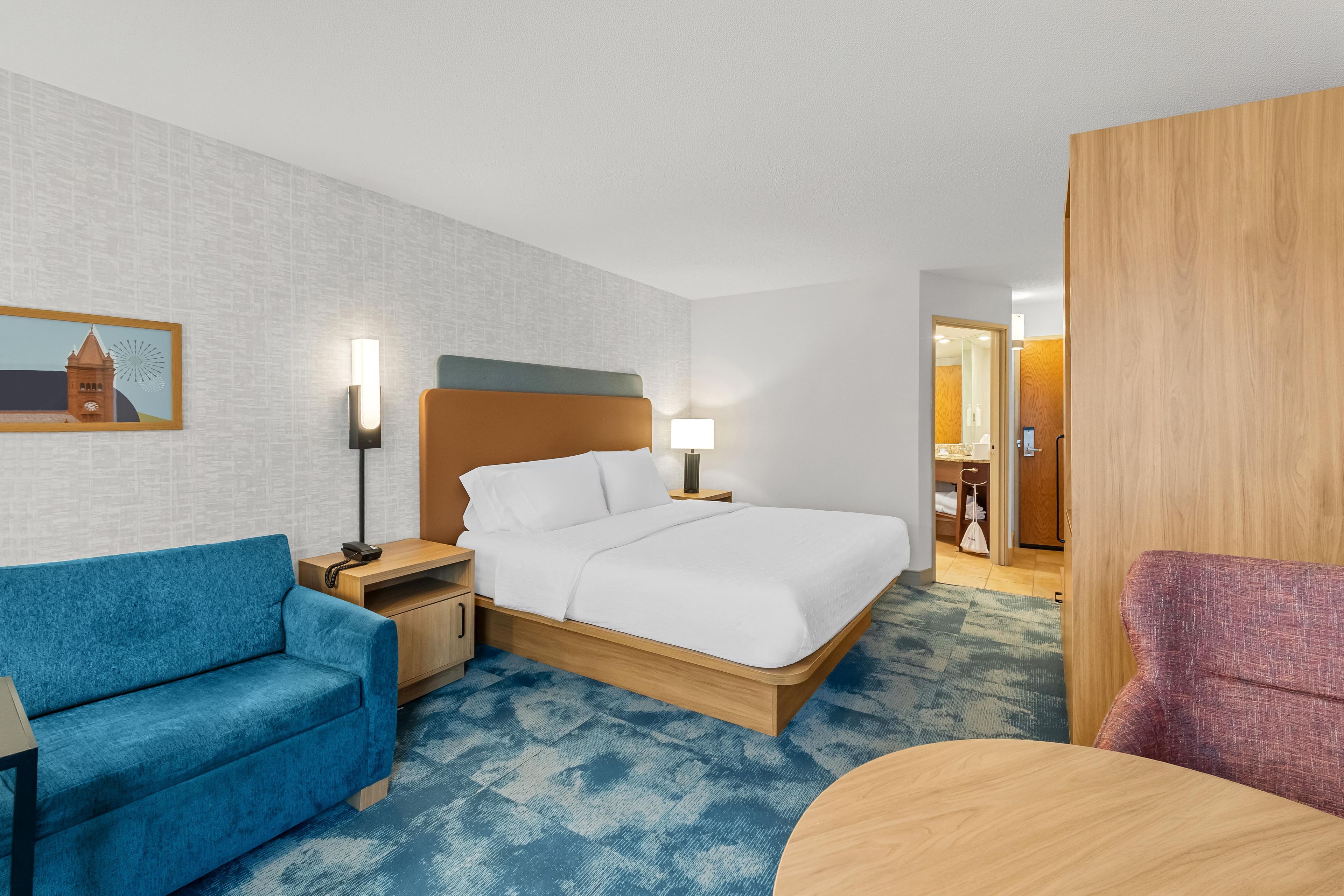 Una habitación de hotel con una cama, un sofá azul, una mesita de noche de madera, una lámpara y una silla de madera.