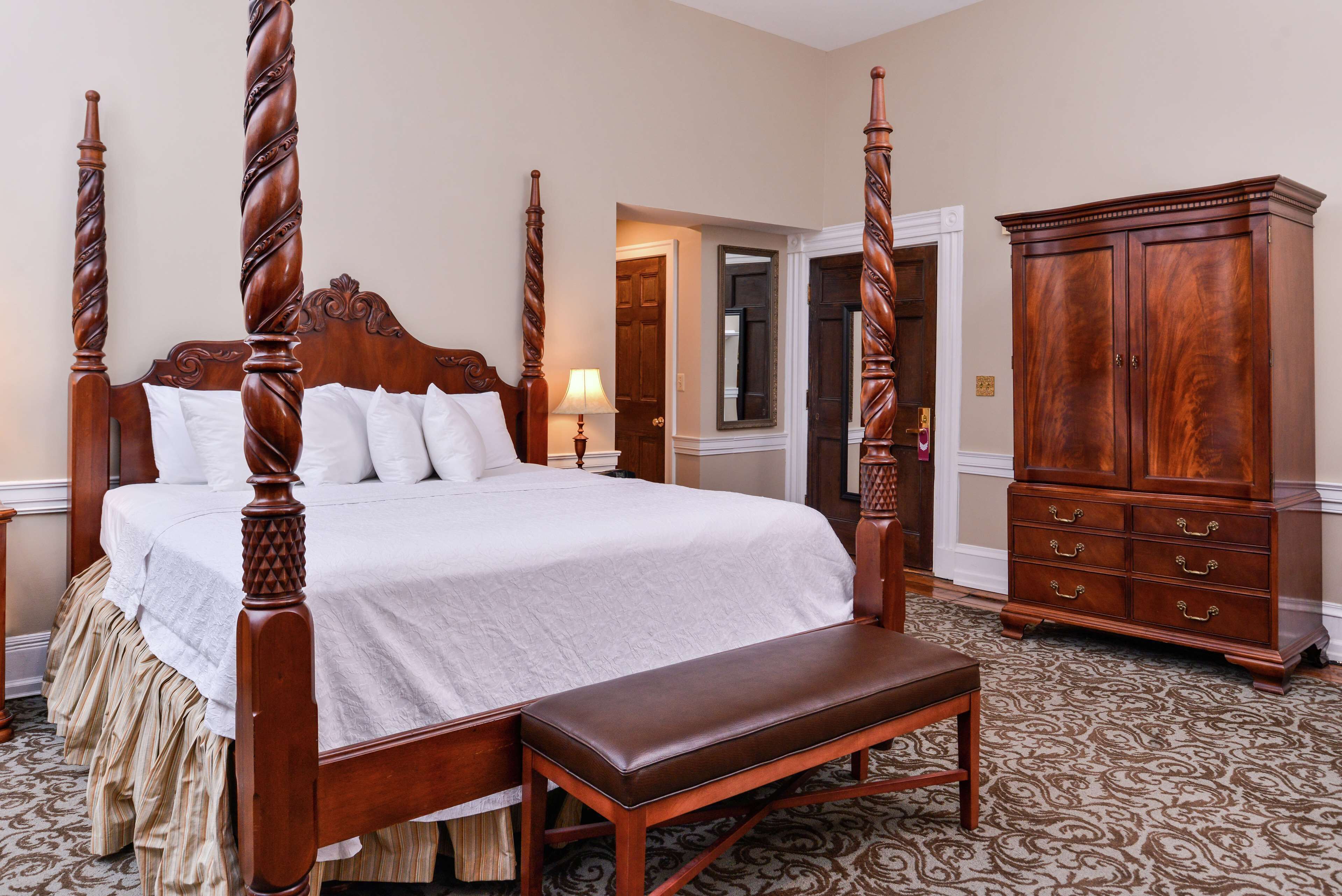 A four-poster bed with a bench, a wooden cabinet, and a patterned carpet in a hotel room.