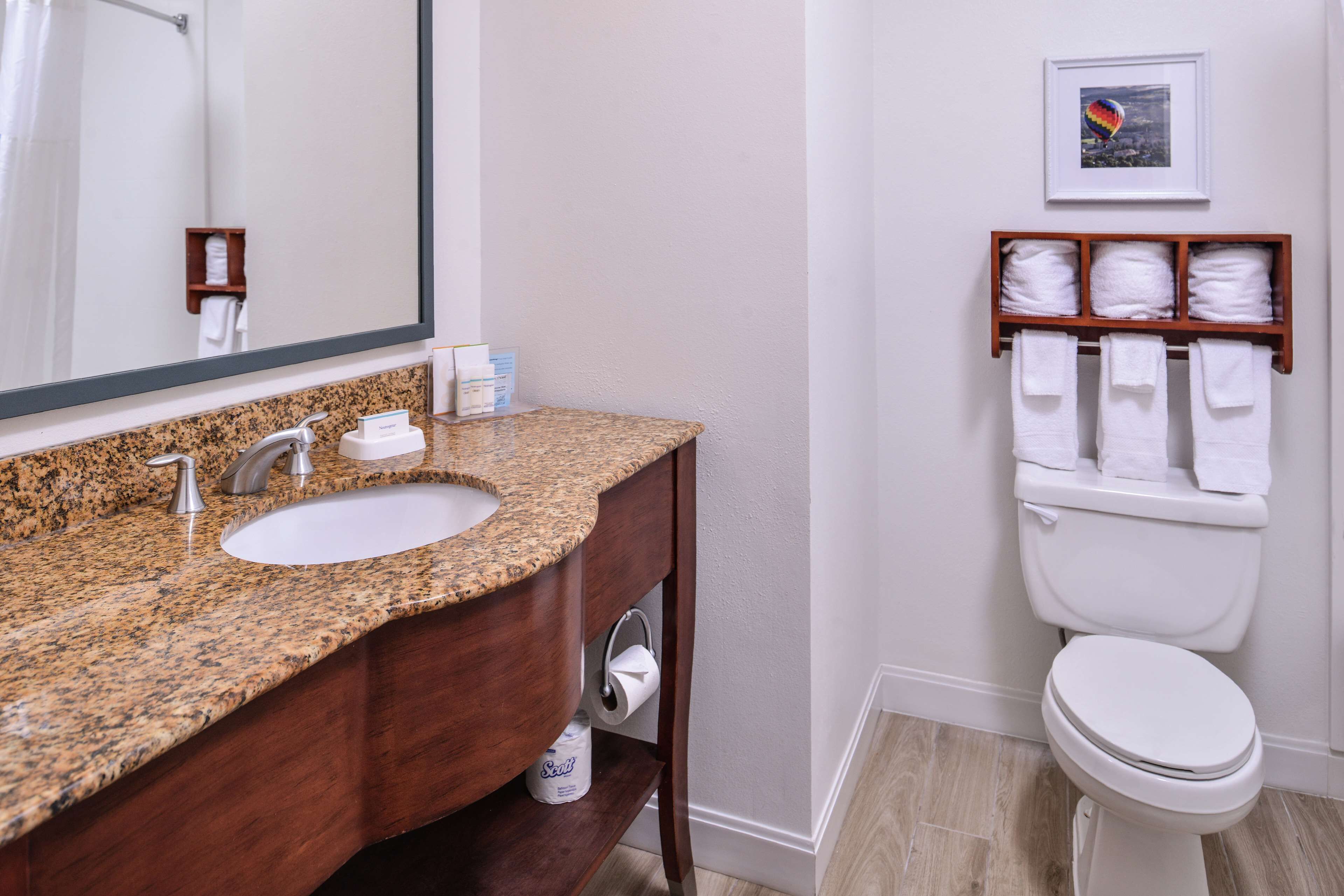 A bathroom with a granite countertop sink, a large mirror, a toilet with a shelf of towels, and a framed picture on the wall.
