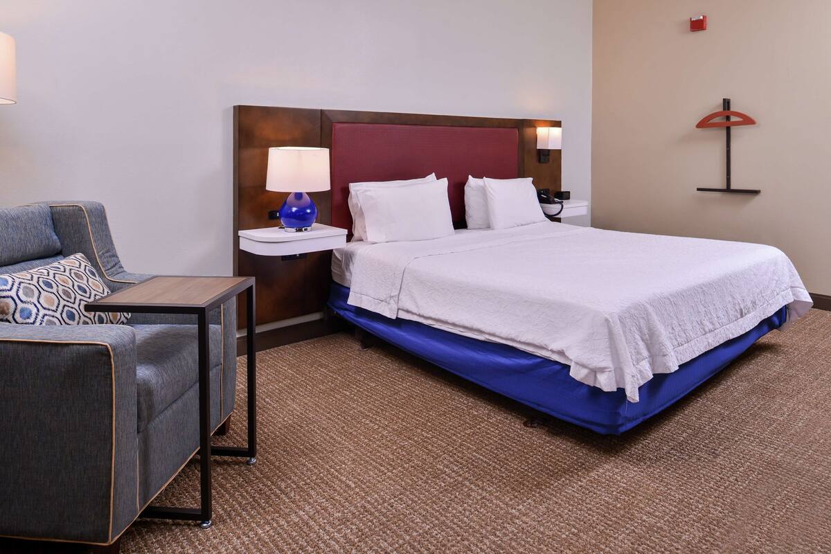 Habitació d'hotel amb un llit, una taula de nit, una làmpada, una cadira i una estant penjada a la paret.