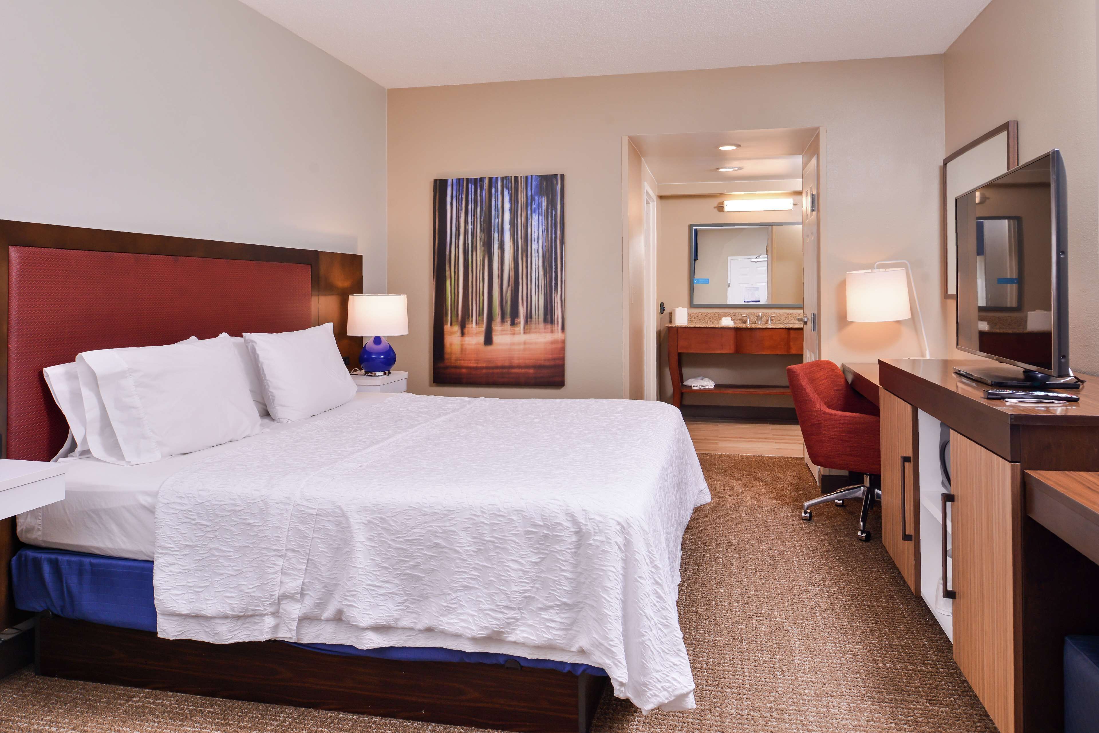 A hotel room with a large bed, a desk with a chair, a mirror, and a painting on the wall.