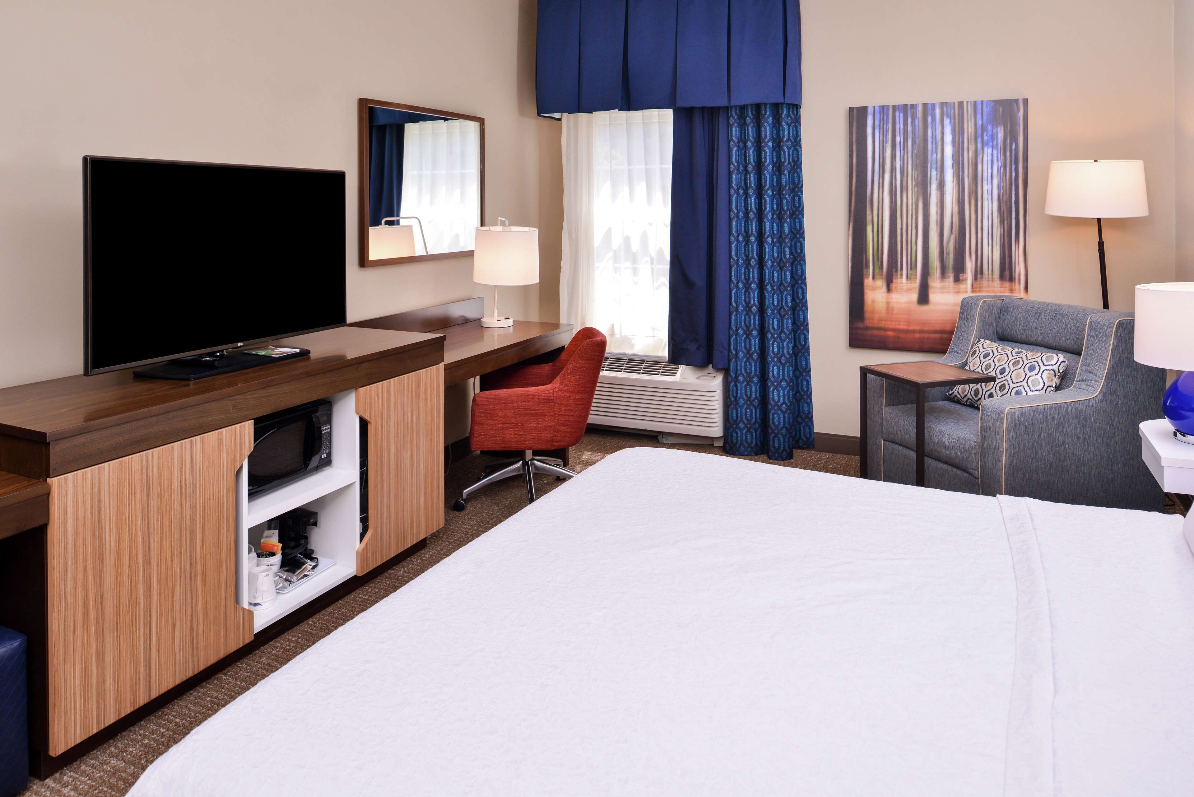 Une chambre d’hôtel avec un grand lit, un bureau doté d’un téléviseur à écran plat, un fauteuil de bureau rouge, un fauteuil gris et une grande fenêtre avec des rideaux bleus.
