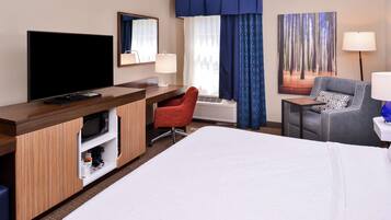 Une chambre d’hôtel avec un grand lit, un bureau doté d’un téléviseur à écran plat, un fauteuil de bureau rouge, un fauteuil gris et une grande fenêtre avec des rideaux bleus.
