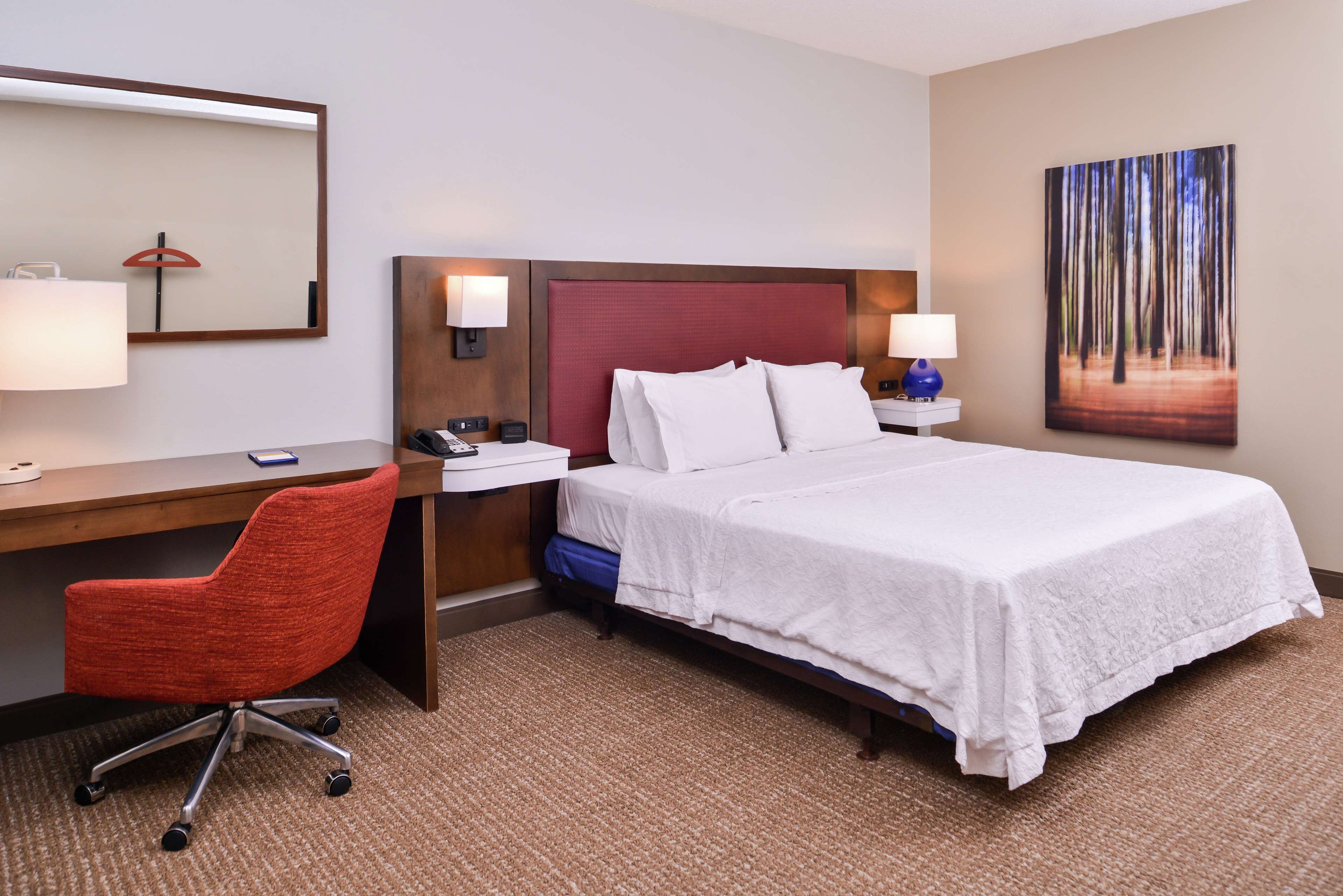 A hotel room with a bed, desk, chair, and a painting on the wall.