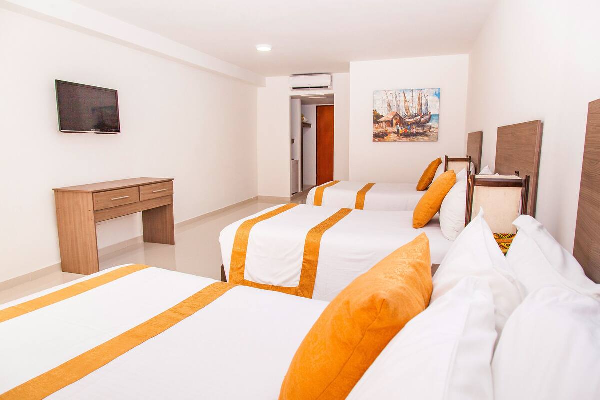 Quarto de hotel com três camas, uma escrivaninha, televisão e ar condicionado.
