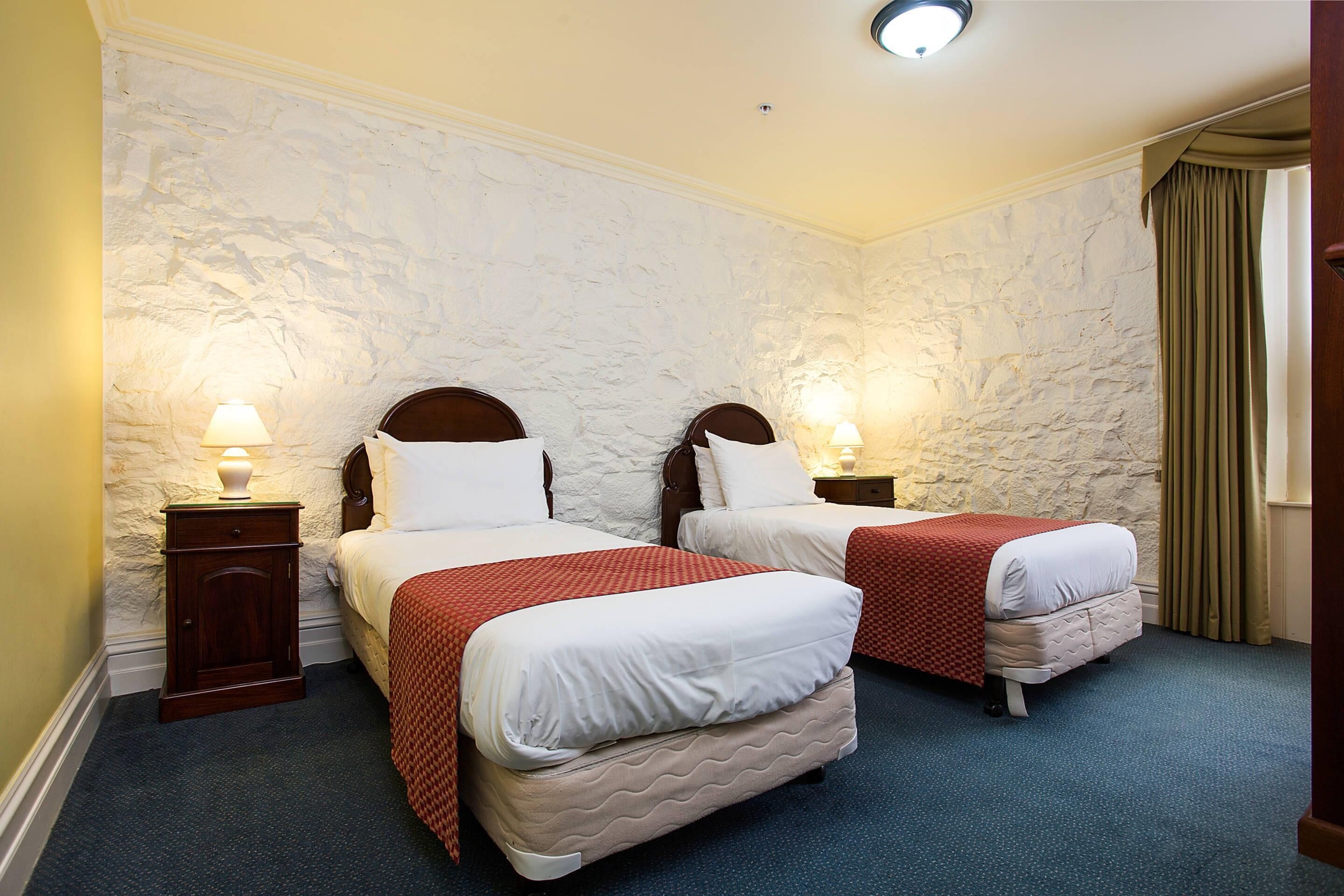 A hotel room with two beds, each with a wooden headboard, white bedding, and a red blanket. There are bedside lamps, a small wooden nightstand, and a stone-patterned wall.
