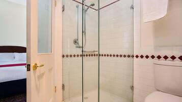 A hotel bathroom with a glass-enclosed shower, a toilet, and a bed with a red-striped blanket.