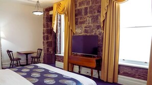 Una habitación de hotel con una cama grande, un escritorio con televisor, una pared de piedra y una ventana con cortinas.