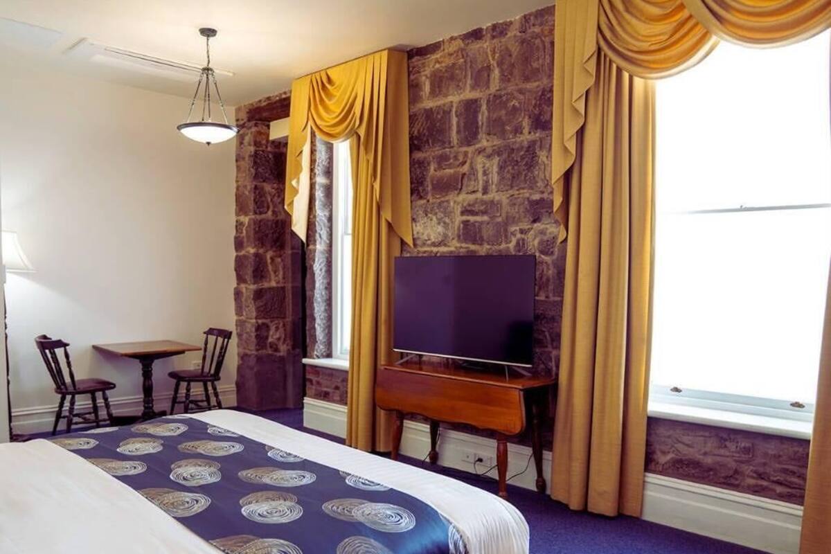 A hotel room with a large bed, a desk with a television, a stone wall, and a window with curtains.