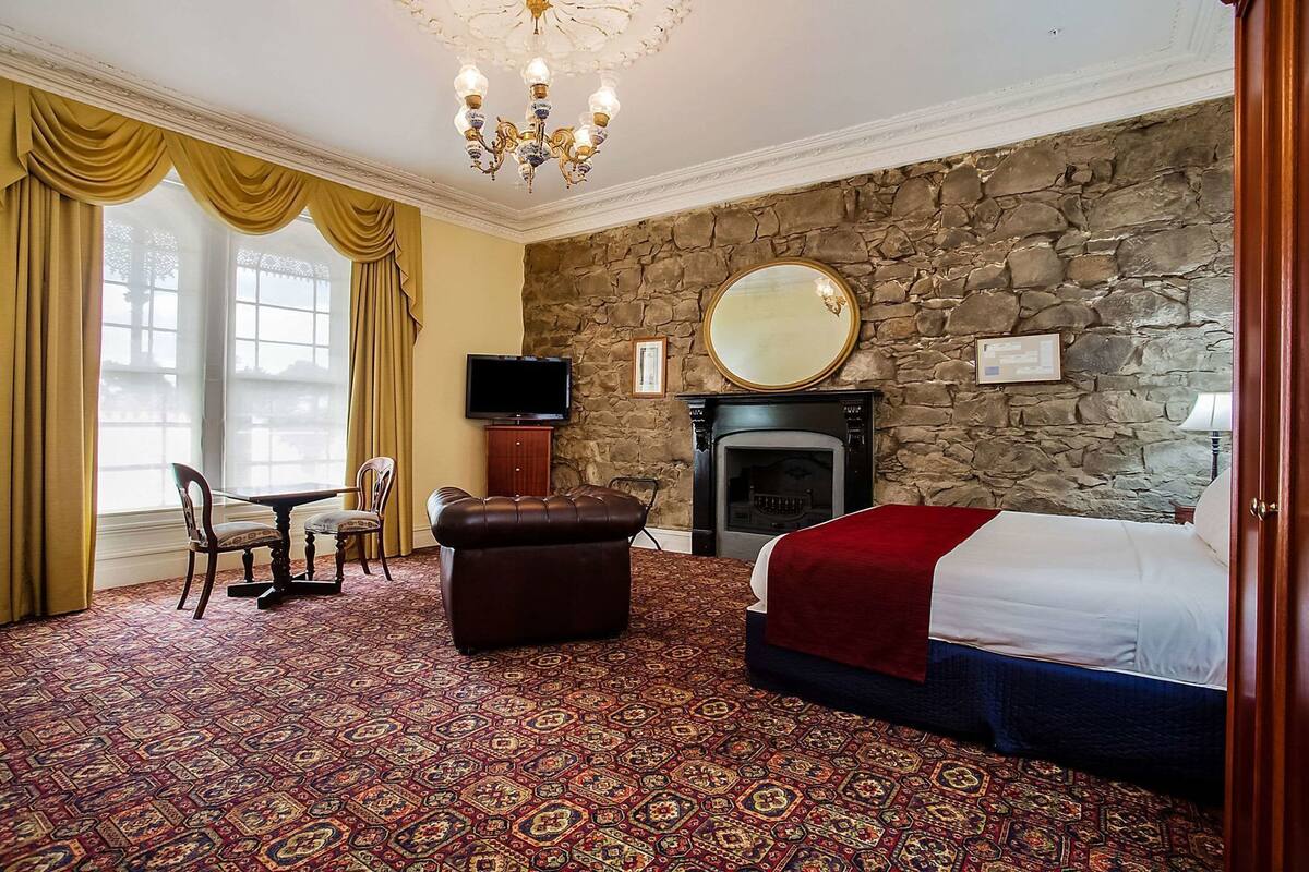 A hotel room with a stone wall, a fireplace, a bed, a television, a desk, and a patterned carpet.
