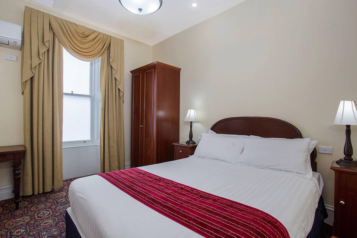 A hotel room with a large bed, a wooden wardrobe, a desk, and a window with curtains.