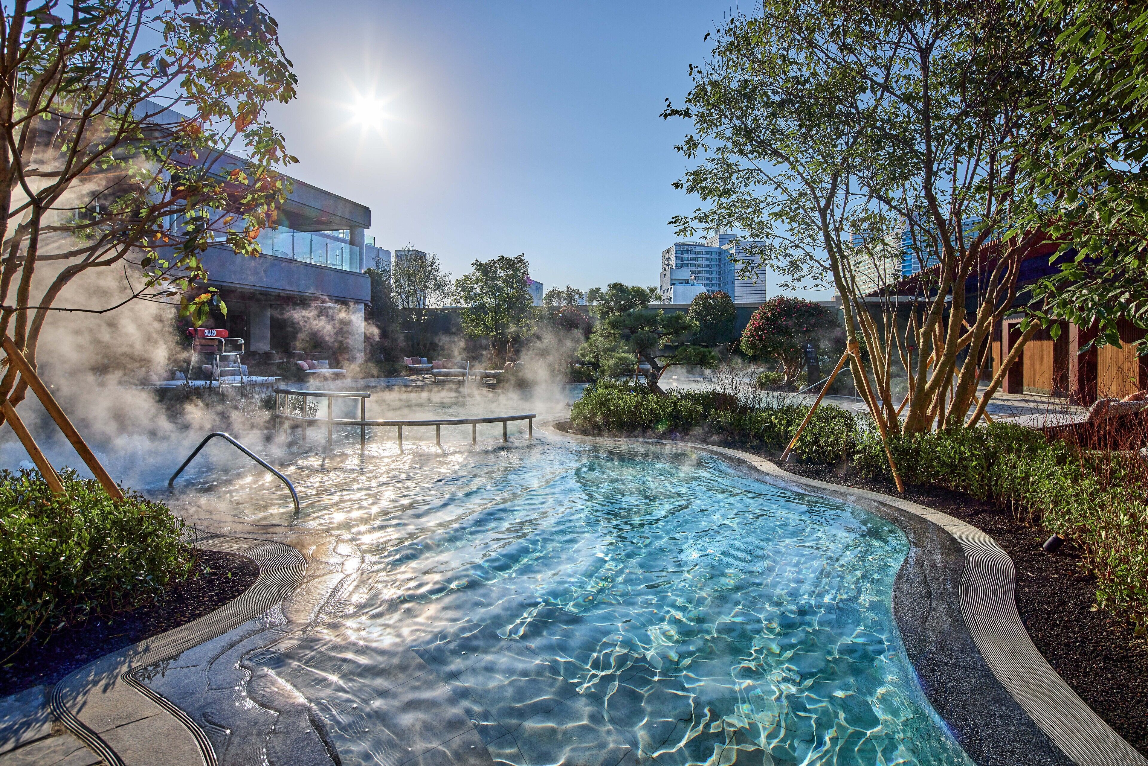 Una piscina con agua cristalina, rodeada de árboles y un edificio moderno al fondo.
