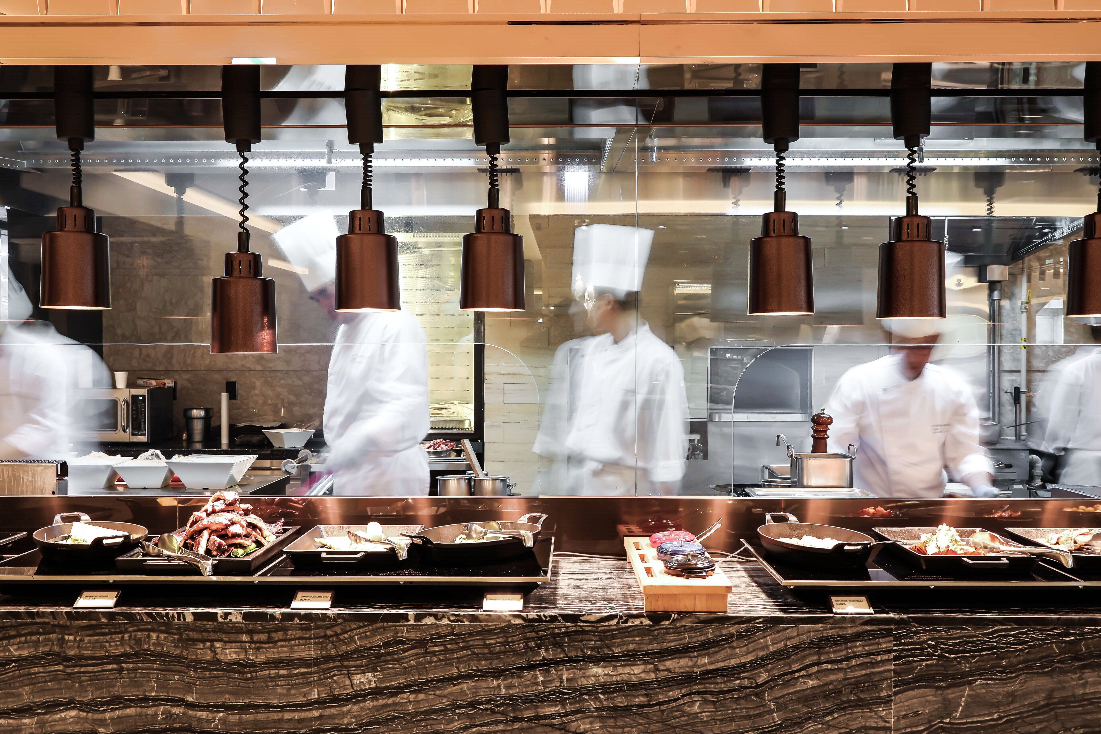 Una cocina con luces colgantes, cocineros con uniformes blancos y distintas estaciones de cocina con bandejas de comida.