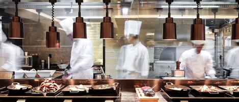 Una cocina con luces colgantes, cocineros con uniformes blancos y distintas estaciones de cocina con bandejas de comida.