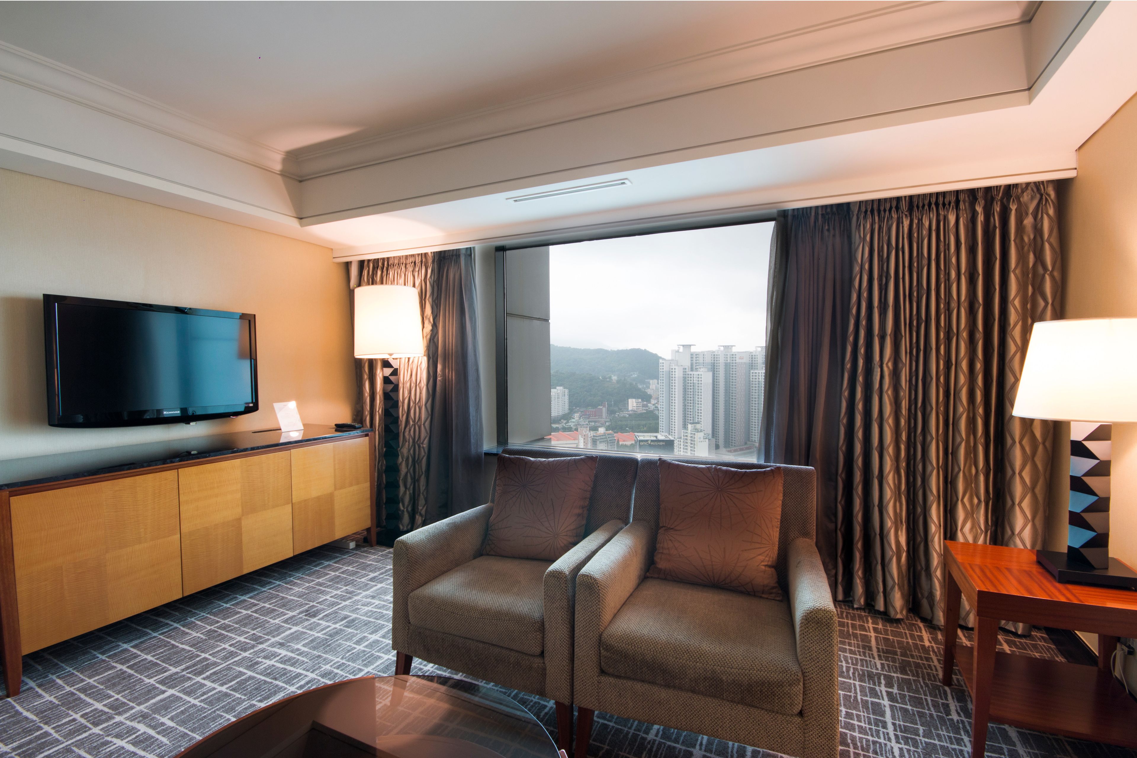 Ein Hotelzimmer mit großem Fenster, Flachbildfernseher, Sofa, Beistelltisch und Lampe.
