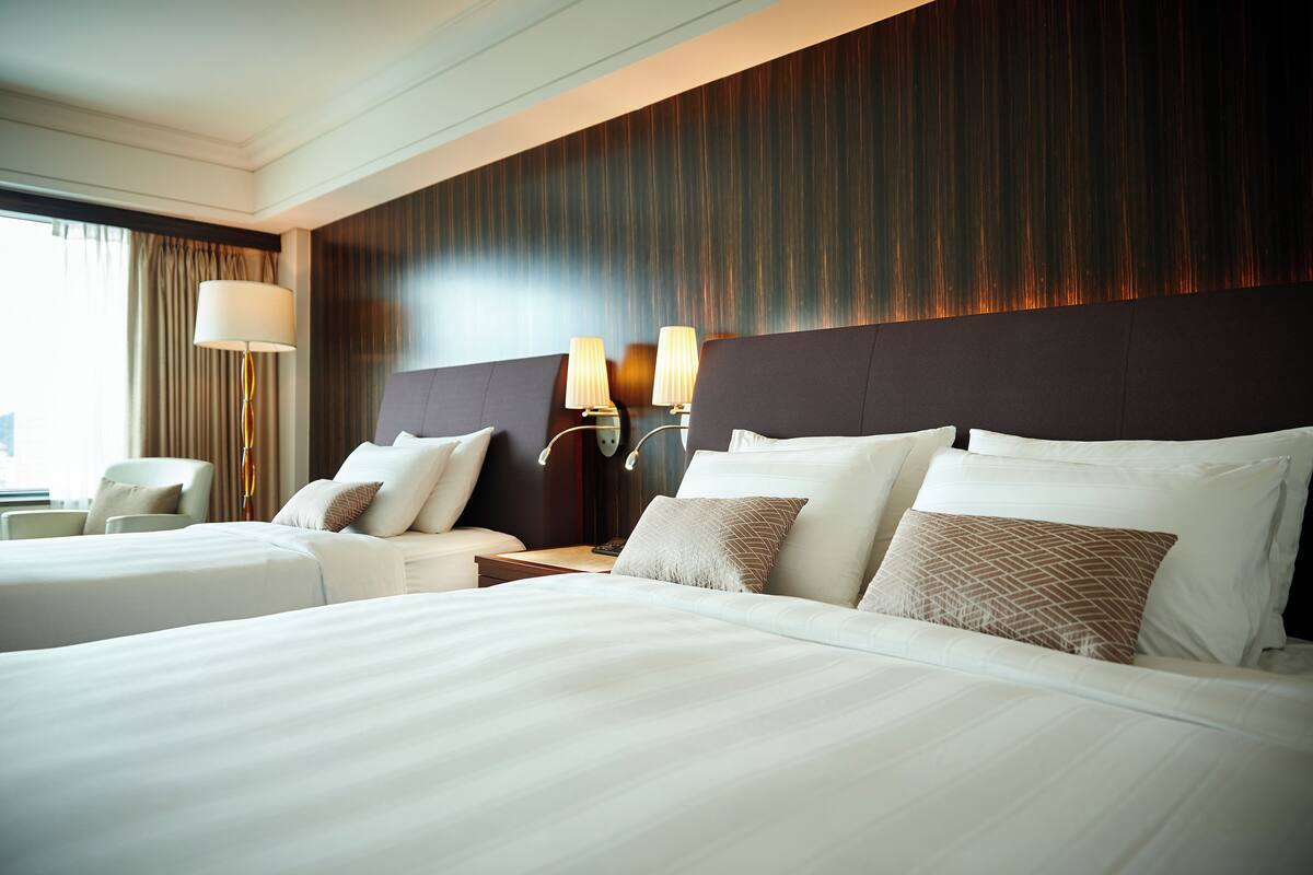 A hotel room with two beds, a nightstand with lamps, and a view of the city through large windows.