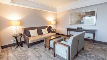 A hotel room with a sofa, armchair, coffee table, lamp, and a desk.