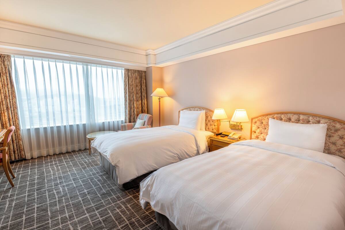 A hotel room with two beds, a desk, a chair, and a large window with curtains.