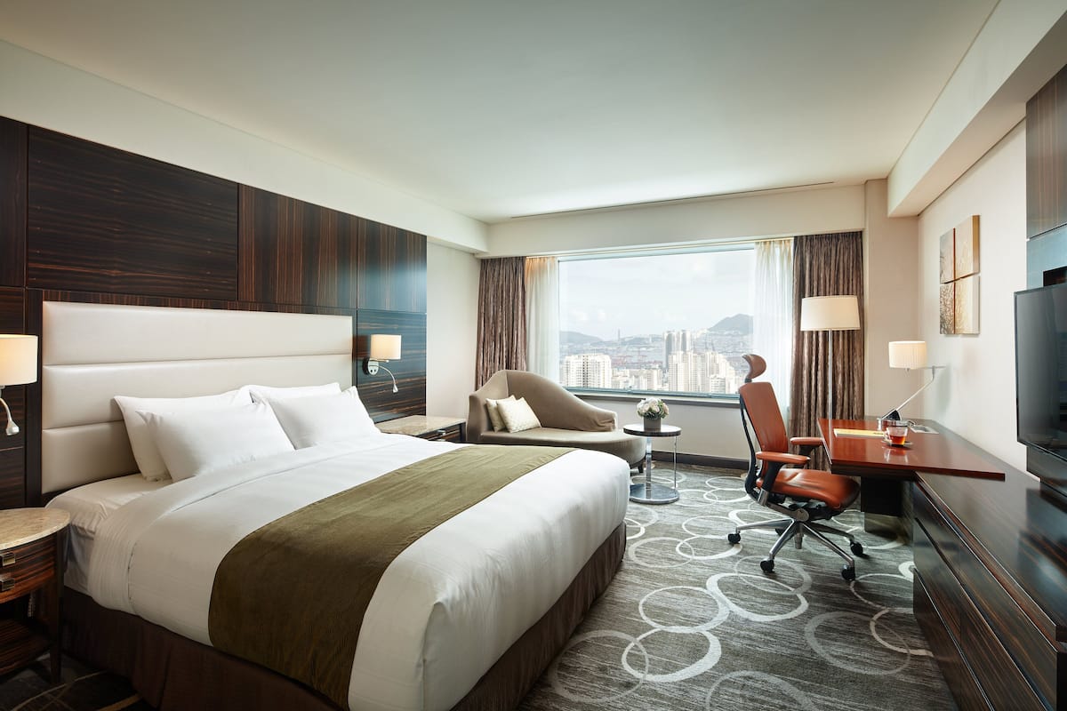 A hotel room with a large bed, a desk, a chair, a TV, and a view of the city.