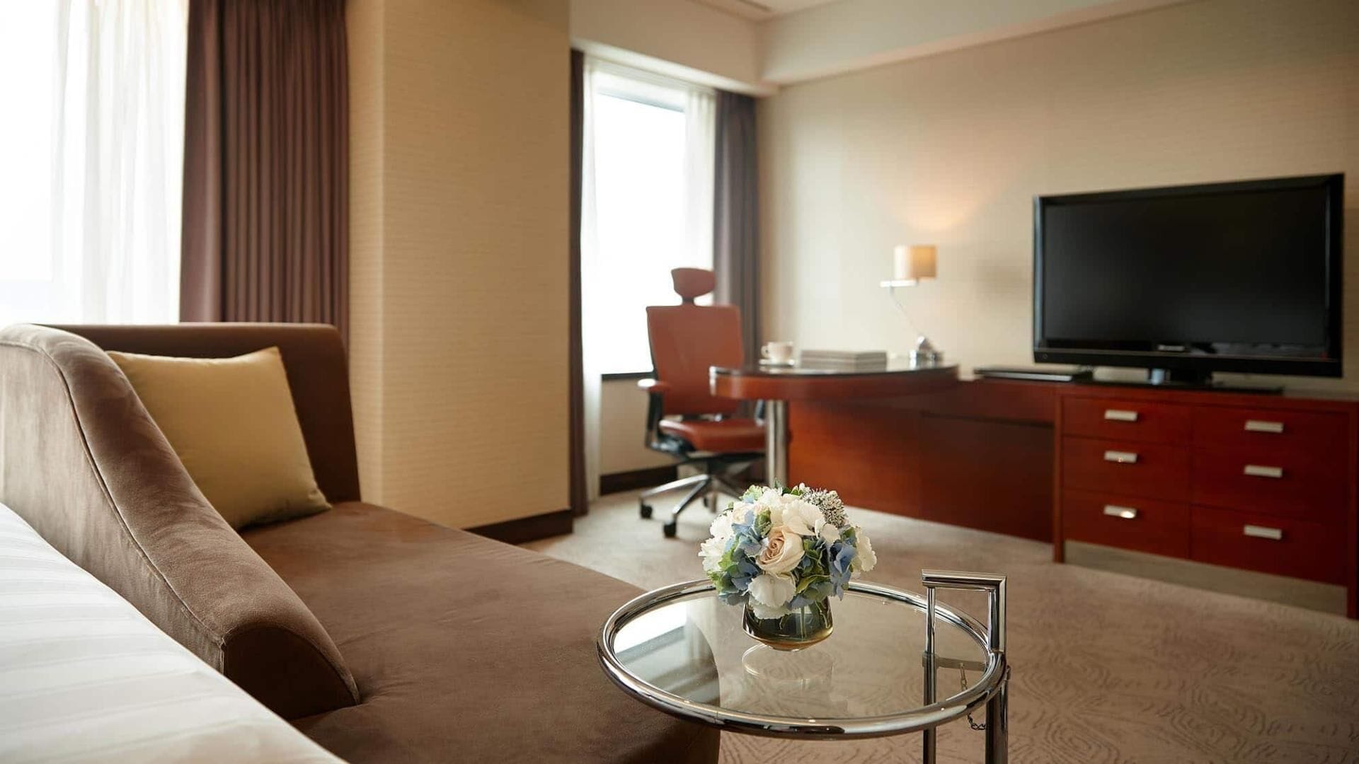 A hotel room with a sofa, a small table with a vase of flowers, a TV, a desk, and a chair.