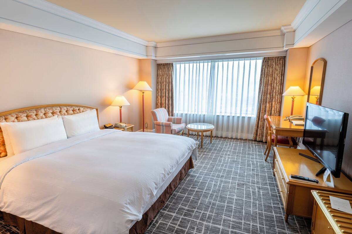 A hotel room with a large bed, a desk with a TV, a chair, a small table, and a window with curtains.