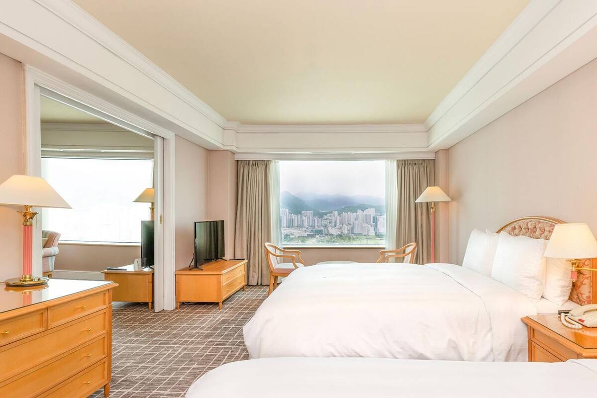 A hotel room with two beds, a desk, a chair, a TV, and a view of a cityscape through the window.