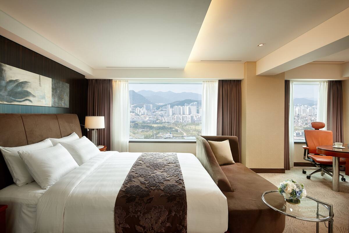 A hotel room with a large bed, a sofa, a desk, and a view of a cityscape.