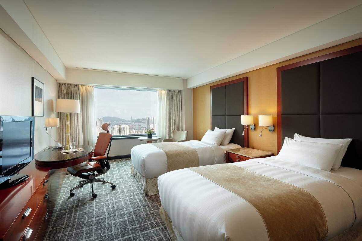 A hotel room with two beds, a desk with a TV, a chair, and a window with a city view.