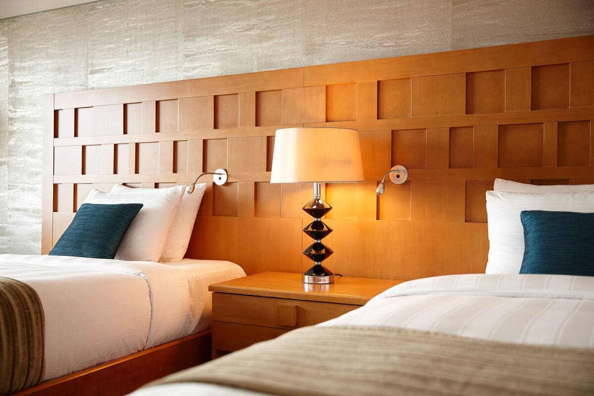 A hotel room with two beds, a wooden headboard, a nightstand with a lamp, and a wall-mounted light fixture.