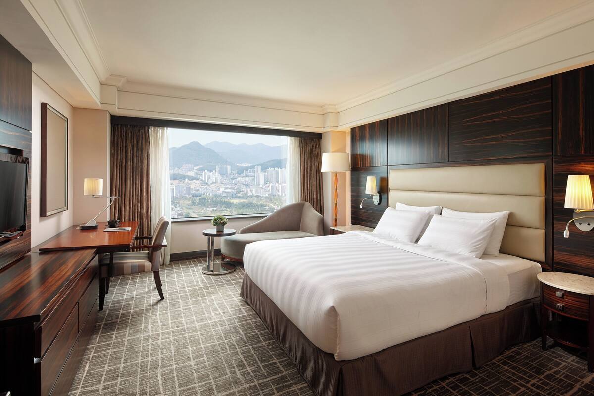A hotel room with a large bed, a desk with a chair, a small table with a lamp, and a view of a cityscape through a large window.