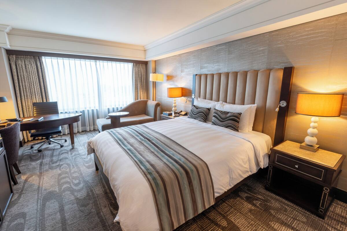 A hotel room with a large bed, a desk, a chair, and a nightstand with lamps.