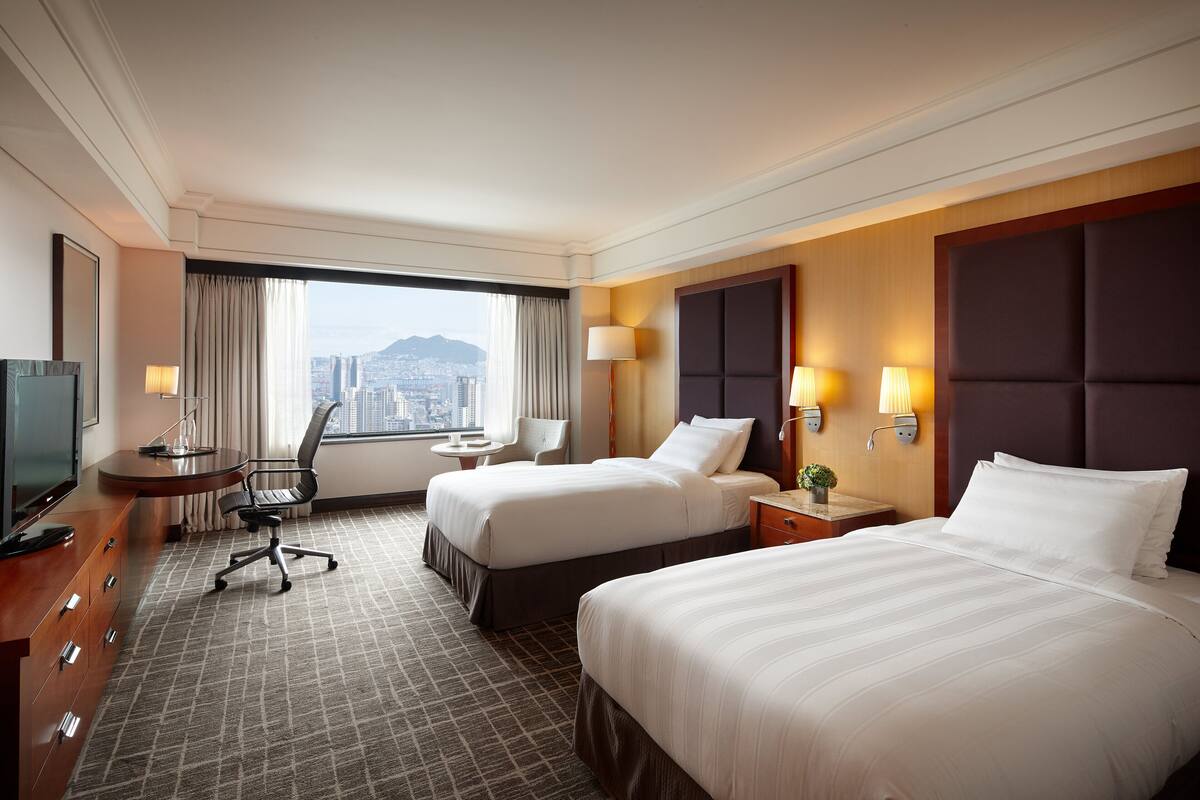 A hotel room with two beds, a desk, a chair, and a TV.