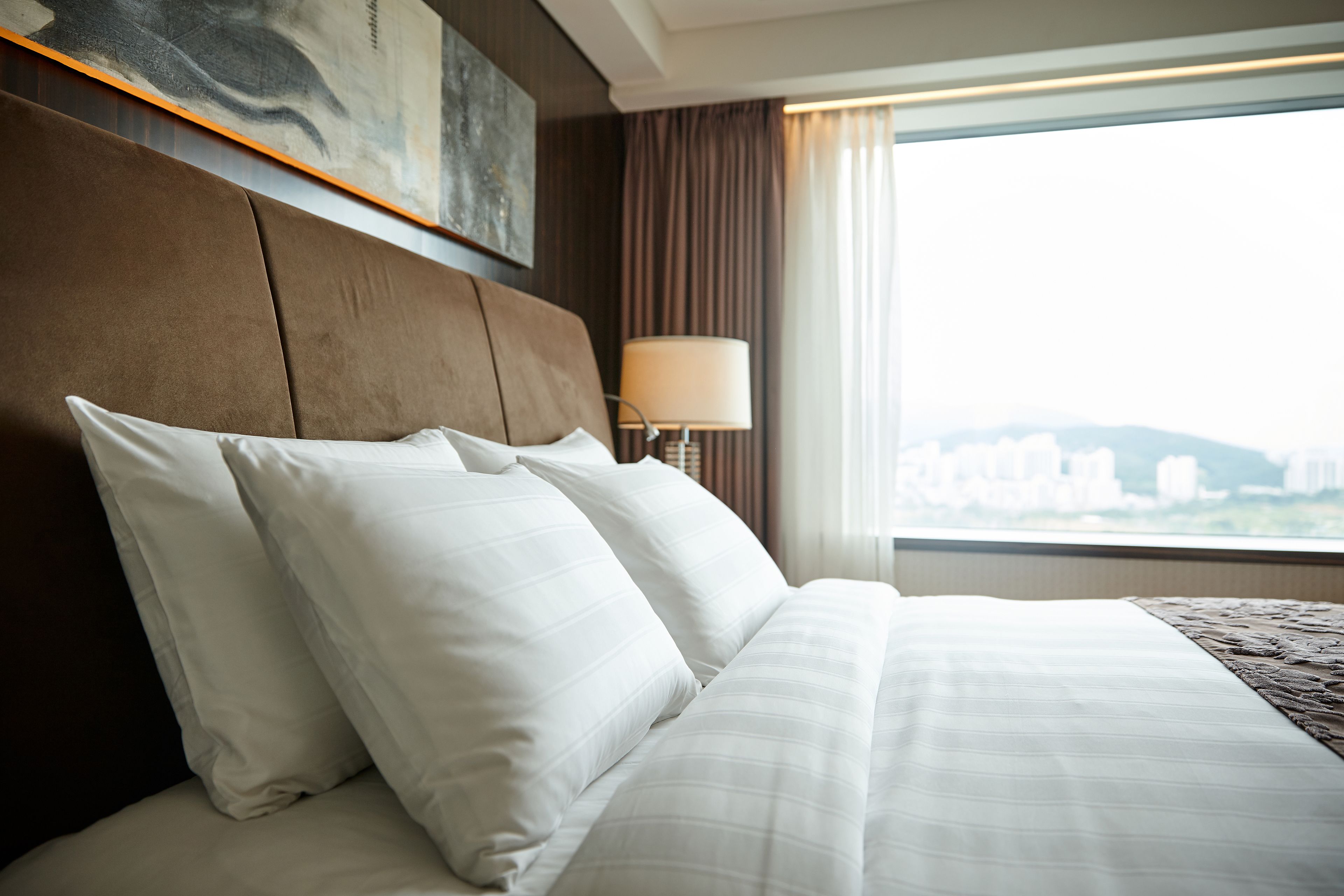 Une chambre d’hôtel avec un grand lit, une tête de lit, une lampe et une vue sur les immeubles par la fenêtre.