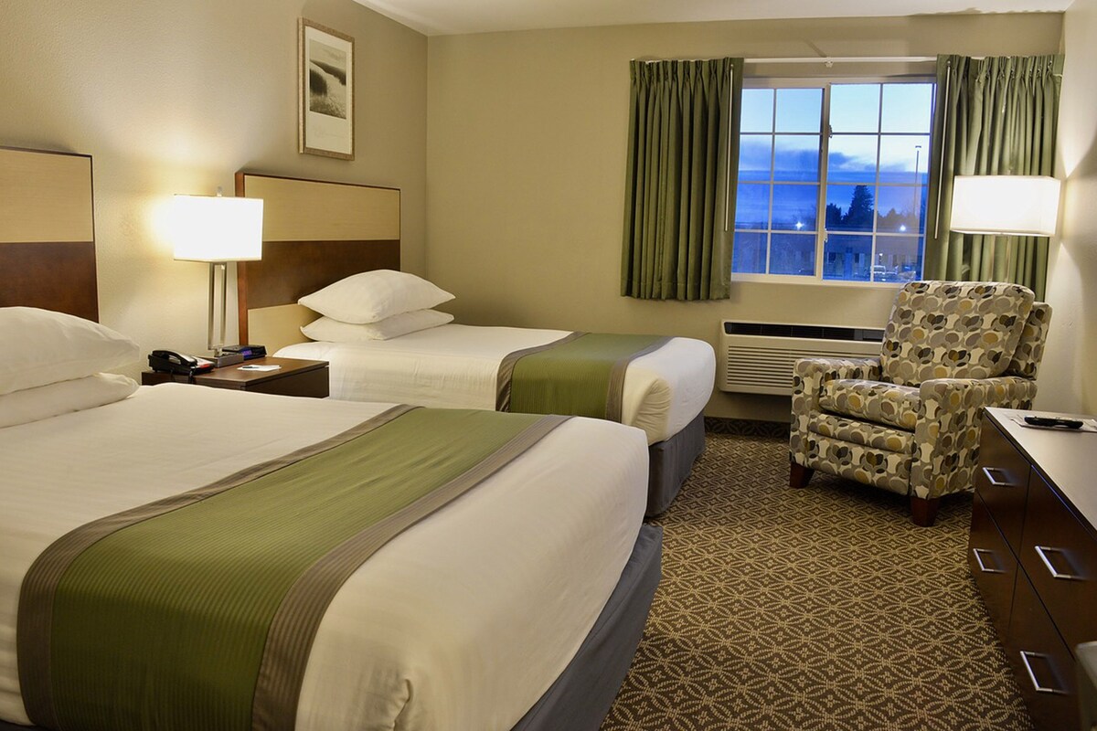 A hotel room with two beds, a desk, a chair, a window with curtains, and a wall-mounted lamp.