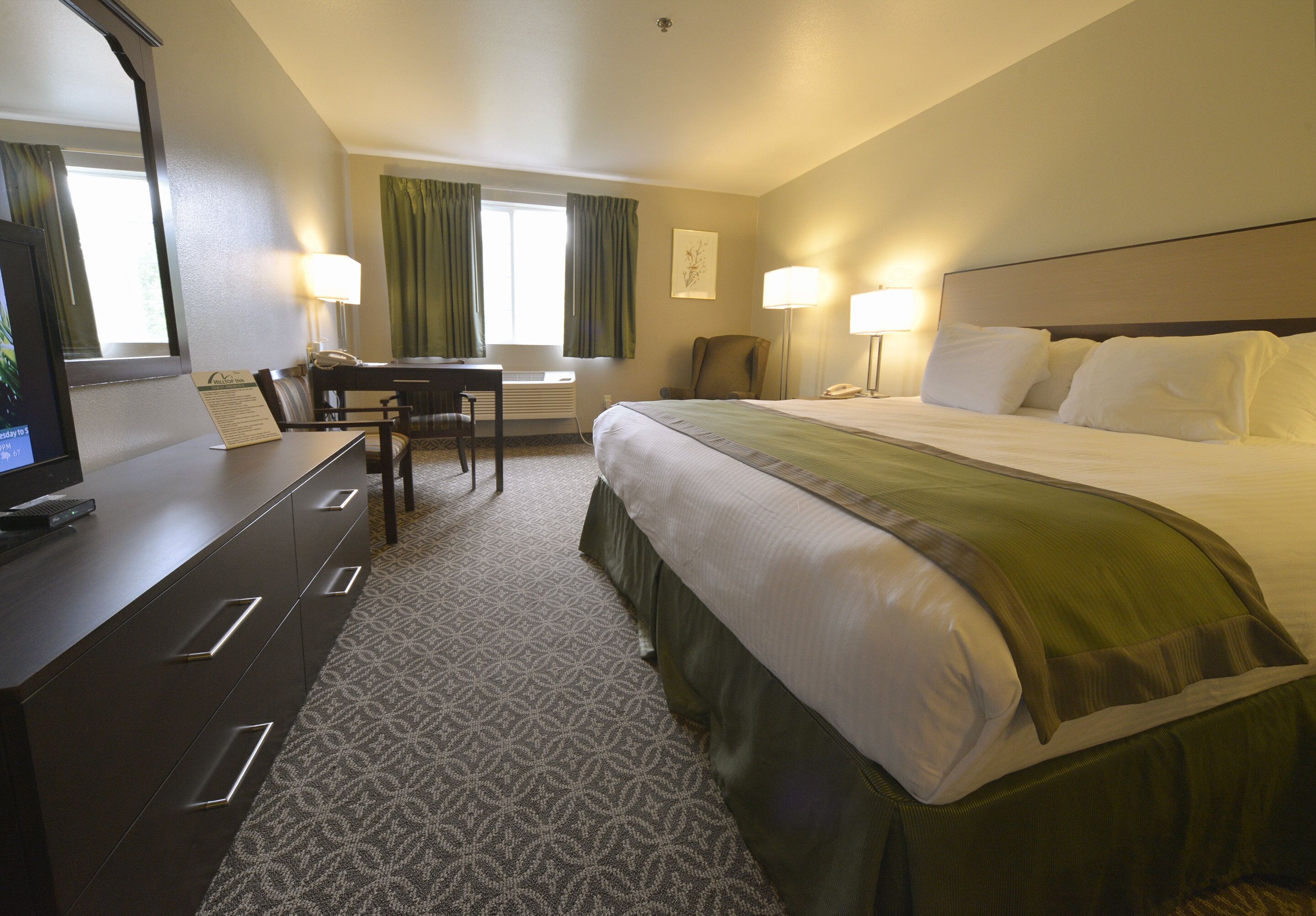 Habitación de hotel con una cama, un escritorio con computadora, una silla, una lámpara y una ventana con cortinas.